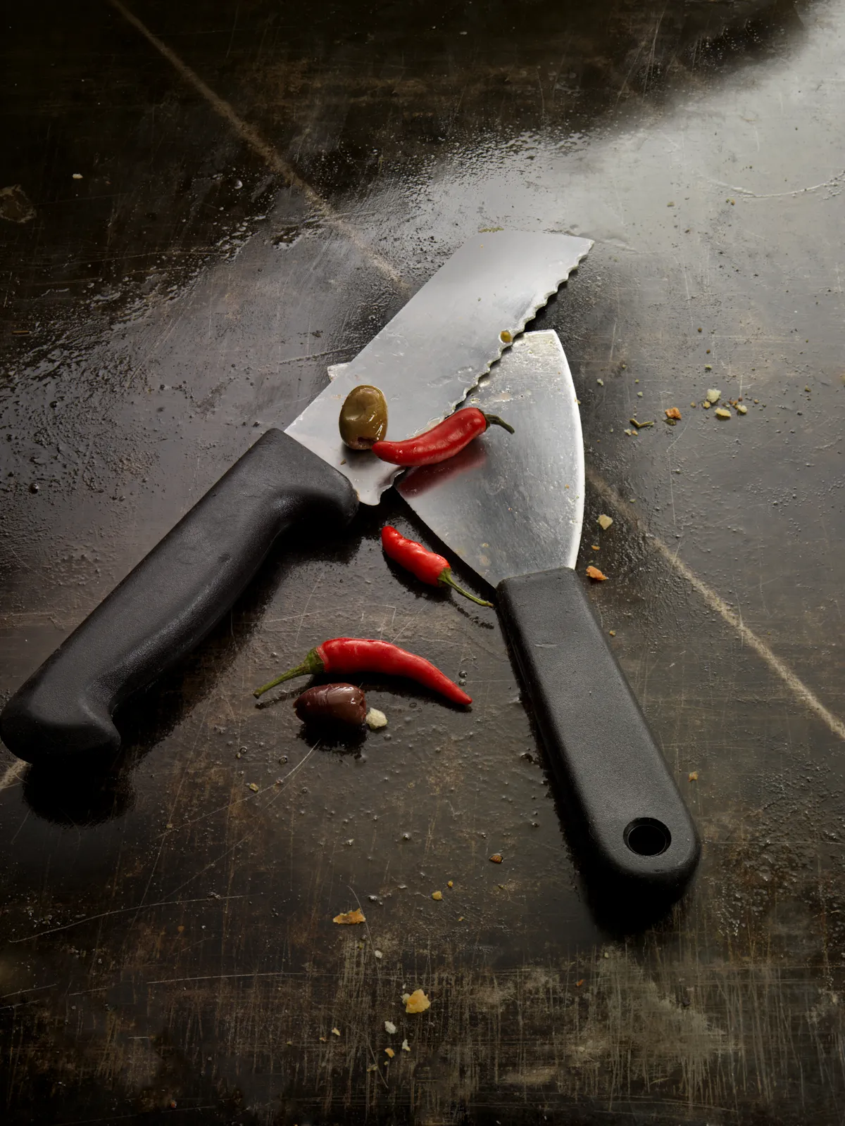 Images qui montrent un couteau et une spatule qui servent à découper les pizzas posées sur une plaque de cuisson, et comme décoration, trois piments et deux olives